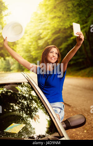 Cheerful woman on summer travel vacation leaning out of a car window. She is holding hat with arms raised and taking selfie. Stock Photo