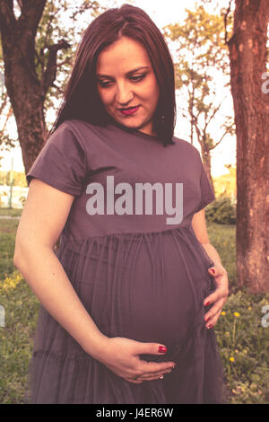 Portrait of happy young pregnant model sitting with crossed legs on grass lawn and looking at her belly with gentle smile. Future mom expecting baby c Stock Photo