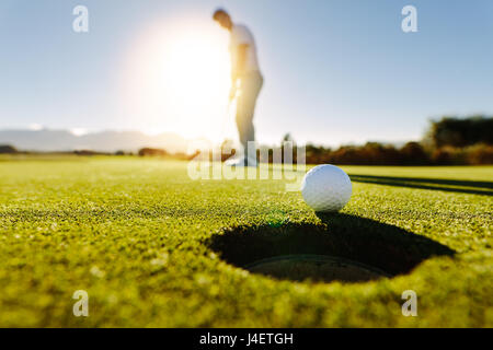 Pro golfer putting golf ball in to the hole. Golf ball by the hole with player in background on a sunny day. Stock Photo