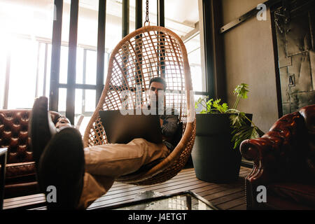 Male executive sitting on a wicker hanging chair in office lounge and looking at laptop. Man relaxing in office lounge working on laptop. Stock Photo