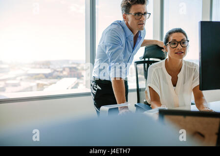 Mature businesswoman sitting at her desk and working on computer with male colleague. Two corporate professionals looking at computer monitor in offic Stock Photo
