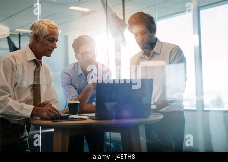 Three male colleagues looking at laptop and discussing. Team of professionals having a standing meeting around table in office. Stock Photo