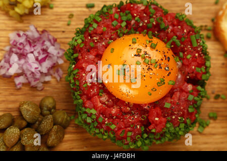 One portion of raw minced beef meat tartare steak with egg yolk, green shallot onions and capers on wooden board, close up, elevated top view, persona Stock Photo