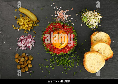 One portion of raw minced beef meat tartare steak with egg yolk, green shallot onions, cucumbers, capers, salt and toasts served on black slate board, Stock Photo