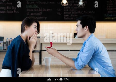 Asian man showing an engagement ring diamond to his amazed girlfriend in a restaurant. Proposal concept Stock Photo