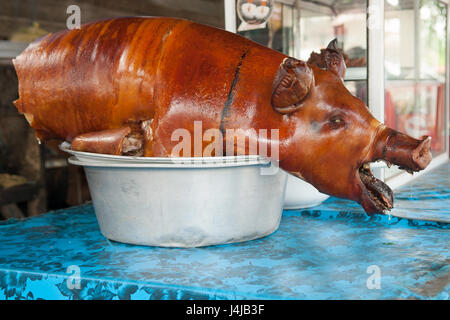 Roasted pink and brown smoked piglet: the bird is cut halfway and placed in a basin on blue counter for sale, the animal's mouth is open. Stock Photo