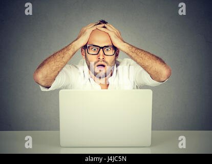 Shocked young man looking at laptop computer anxious with open mouth and big eyes hands on head in disbelief. Human emotion reaction Stock Photo