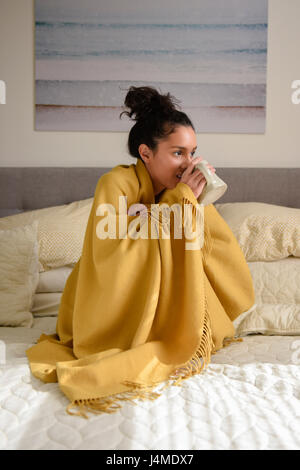 Hispanic woman sitting on bed wrapped in blanket drinking coffee Stock Photo