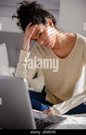 Frustrated Hispanic woman sitting on bed using laptop Stock Photo