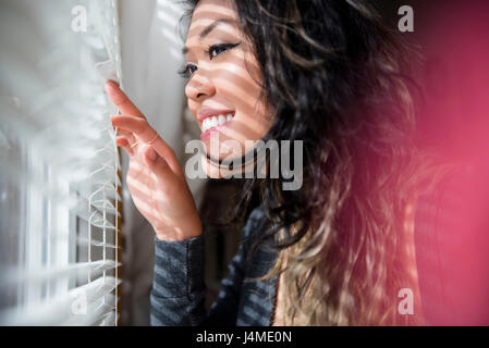 Mixed Race woman peeking out window behind blinds Stock Photo