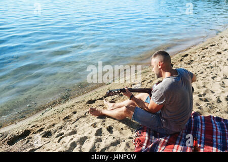 Young man playing guitar while relaxing on sandy beach Stock Photo