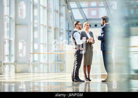 Group of modern business people chatting during coffee break  standing in sunlit glass hall of office building Stock Photo
