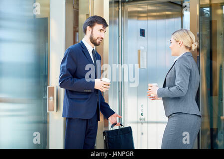 Portrait of two business colleagues dressed in formalwear meeting and talking by elevator on way to work, both holding coffee cups Stock Photo