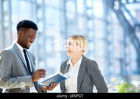 Portrait of two young business colleagues, one of them African,  discussing work smiling cheerfully  standing in glass hall of modern office building Stock Photo