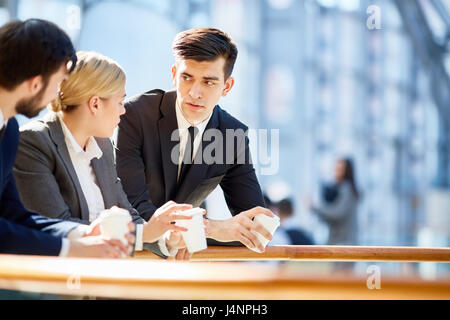 Portrait of three business people, one woman and two men, discussing work at break  in modern office building leaning on railing and holding coffee cu Stock Photo