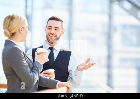 Portrait of two successful business people, man and woman, smiling and talking talking during break in gass hall of modern building, both holding coff Stock Photo