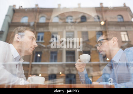 Side view portrait of two businessmen sitting at opposite sides of table in cafe discussing deal during meeting, shot from behind glass window Stock Photo