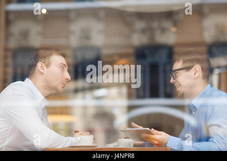 Side view portrait of two men meeting in cafe sitting on opposite sides of table and discussing work, shot from behind glass window Stock Photo