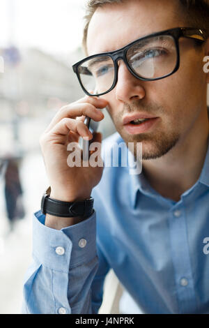 Portrait of young businessman wearing big black glasses discussing work speaking by mobile phone at window in cafe Stock Photo