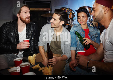 Group of modern young people enjoying party in small night club, sitting on stage, chatting and drinking beer Stock Photo