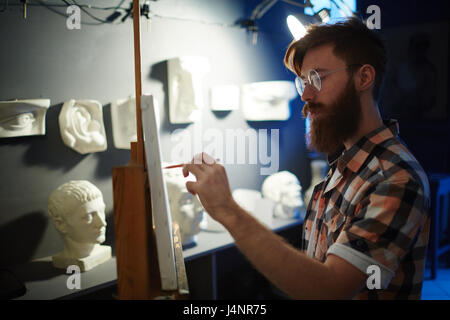 Portrait of inspired young red-haired man painting picture of plaster heads in art studio, looking focused and concentrated Stock Photo