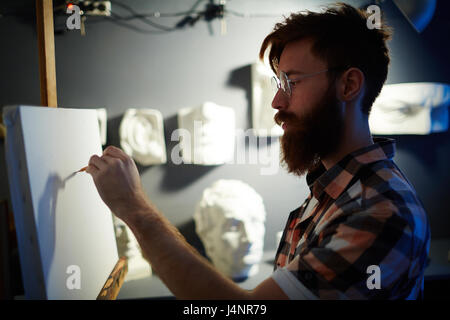 Portrait of modern bearded artist drawing first lines of picture on blank white canvas in dim lighting Stock Photo