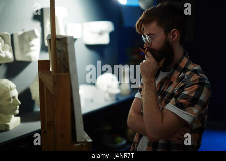 Side view portrait of contemporary bearded artist looking at his panting on canvas deep in thought while working in art studio Stock Photo