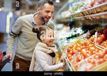 Portrait of cute little girl with dad leaning over vegetable counter choosing fresh ripe tomatoes and other vegetables in supermarket Stock Photo