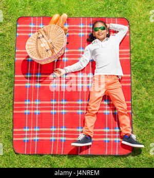Top view portrait of African boy in sunglasses laying on red checkered plaid next to the picnic basket Stock Photo
