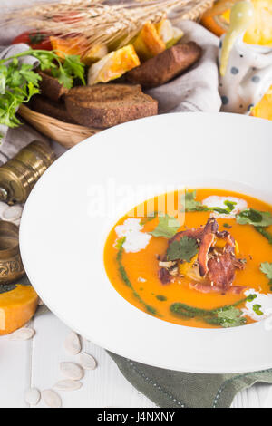 pumpkin soup with bacon in a rustic style on the table. Bread and greens in the background Stock Photo