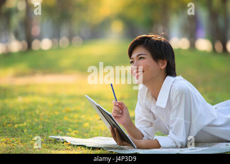 young woman lying on green grass park with pencil and note book in hand thinking something ,project,dream,hope,solution in future Stock Photo