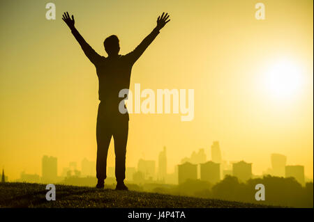 Young man spreads his arms wide in silhouette against a golden sunrise over the city skyline Stock Photo