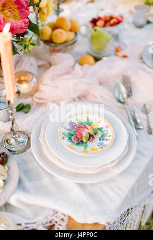 picnic, food, holiday concept - holiday decorated table with white tablecloth, stack of white plates with floral patterns, two acorns on top, burning candle in candlestick, flowers, plates with fruits Stock Photo