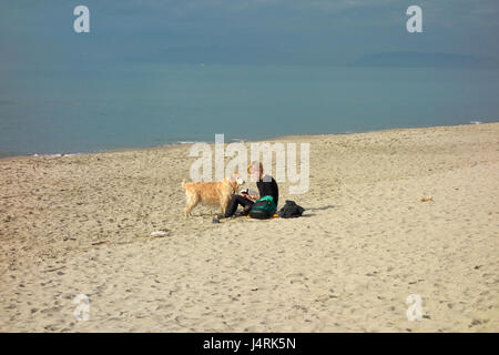 Young blonde woman reading a book with her dog (Golden Retriever) looking at her on the sand beach of Viareggio, Tuscany, Italy, Europe Stock Photo