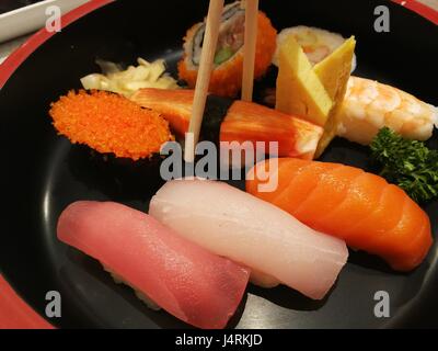 Hand using chopsticks pick Sushi and Sashimi rolls on a black stone slatter. Fresh made Sushi set with salmon, prawns, wasabi and ginger. Stock Photo