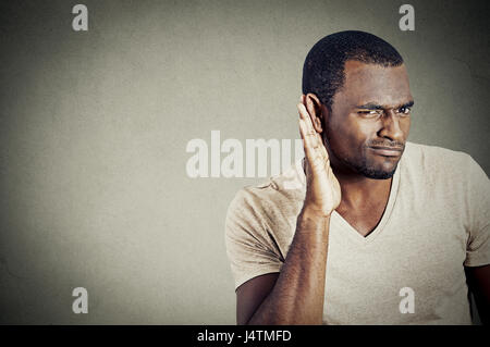 Closeup portrait of handsome young guy trying to secretly listen in on a conversation and not pleased by what he hears isolated on gray wall backgroun Stock Photo