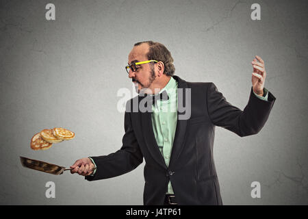 Happy smiling man tossing pancakes on frying pan isolated on grey wall background. Positive face expression emotion, Kitchen fun concept Stock Photo