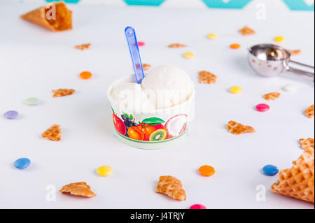 Close up Vanilla Ice-cream in a paper cup on the white background with colorful candies, pieces of waffles, ice cream spoon and empty ice cream cone.  Stock Photo