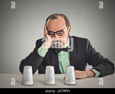 Desperate sad middle aged business man with glasses sitting at desk looking down playing a conjuring trick game isolated grey wall office background.  Stock Photo