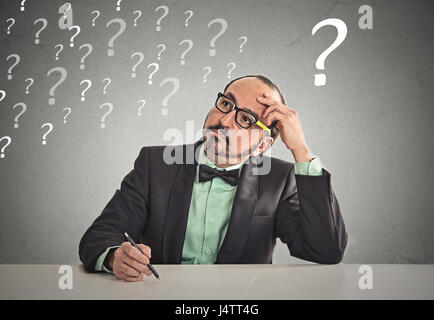 Confused puzzled business man sitting at table scratching his head thinking has many questions isolated office grey wall background. Human face expres Stock Photo