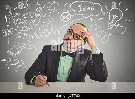 Thoughtful man sitting at table scratching his head thinking ready to write something isolated office grey wall background. Human face expression emot Stock Photo