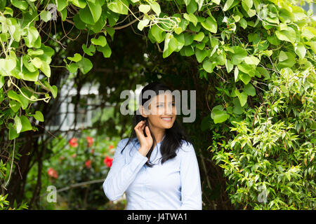 Closeup portrait of confident smiling happy pretty young woman in blue shirt enjoying a fresh new day, isolated background of green shrubs and flowers Stock Photo