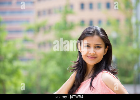 Closeup portrait of confident smiling happy pretty young woman in pink dress, isolated background of blurred trees, buildings. Positive human emotion  Stock Photo