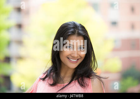 Closeup portrait of confident smiling happy pretty young woman in pink dress, isolated background of blurred trees, buildings. Positive human emotion  Stock Photo
