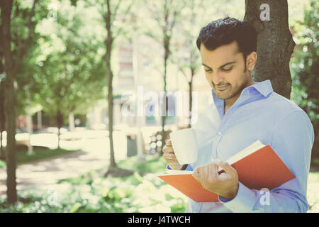 Closeup portrait young business man reading red book and drinking from mug, standing outside on sunny day, isolated green trees background Stock Photo