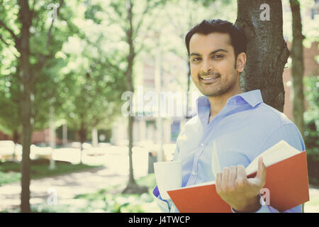Closeup portrait young business man reading red book and drinking from mug, standing outside on sunny day, isolated green trees background Stock Photo