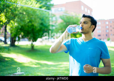 Closeup portrait of young guy in blue shirt drinking water from crystal clear bottle on a hot, sunny day, isolated green trees and building background