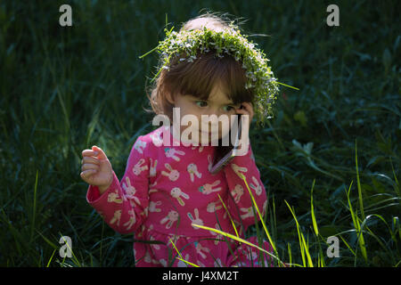 Beautiful little blonde girl with flowers head wreath and talking on her cell phone in the park Stock Photo