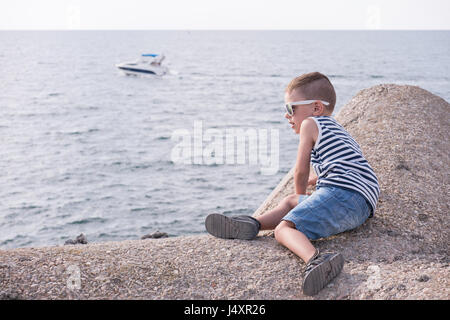 Little boy with sunglasses and striped vest sitting on concrete breakwater in front of the sea with floating boat Stock Photo