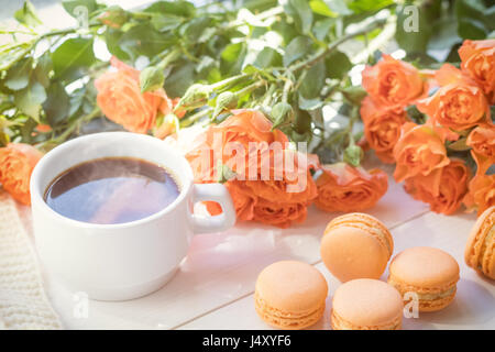 Orange mango or citrous macaroons, cup of coffee and fresh little roses on light wooden background. Sunlight. Coloring and processing photo with light Stock Photo